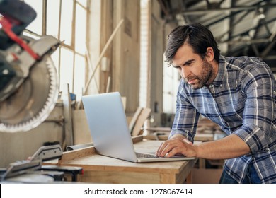 Focused Woodworker Leaning On A Workbench By A Mitre Saw Working Online With A Laptop In His Woodworking Studio