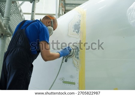 A focused worker uses a sanding tool to smooth an aircraft surface inside a hangar. Protective gear is worn for safety while preparing the plane for a fresh coat of paint.