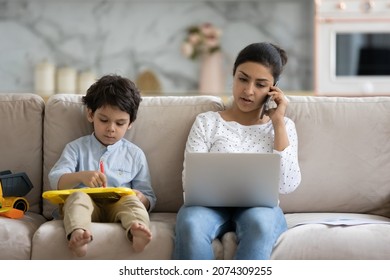Focused working Indian mom talking on mobile phone, holding laptop on couch, watching little son, kid playing with toy drawing board. Motherhood, career, distance job, quarantine concept