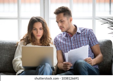 Focused worried couple paying bills online on laptop with documents sitting together on sofa at home, serious confused man and woman planning budget expenses, young family having debt loan problems