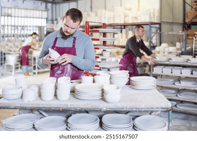 Focused young adult bearded pottery expert in maroon apron meticulously inspecting quality of freshly produced handmade plate in artisanal ceramics studio.. - Powered by Shutterstock