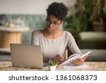 Focused young african american businesswoman or student looking at laptop holding book learning, serious black woman working or studying with computer doing research or preparing for exam online