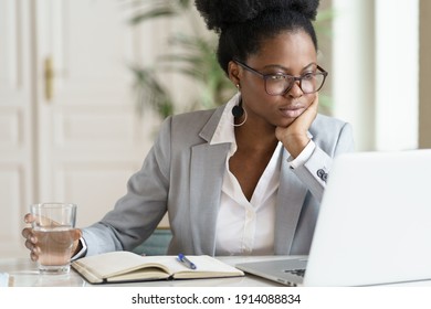 Focused young African American businesswoman or student in blazer wear glasses working at laptop at home office, holding glass of water, looking at screen computer, reading interesting article online.