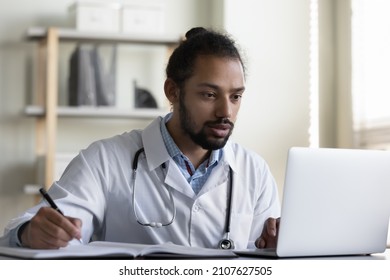 Focused Young African American General Practitioner Doctor Physician Therapist Handwriting Notes In Paper Book, Working On Laptop In Modern Clinic, Managing Appointments Or Consulting Online.