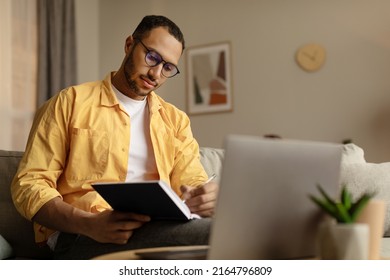 Focused Young African American Man Using Laptop, Working Online, Taking Notes During Business Meeting At Home, Free Space. Millennial Black Guy Learning Online, Writing Down Info