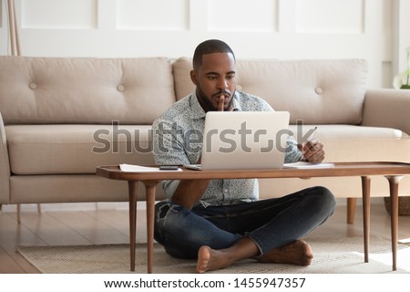 Focused young african american man studying or working on laptop sit on floor in living room, serious black male student freelancer using looking at computer writing notes doing research at home