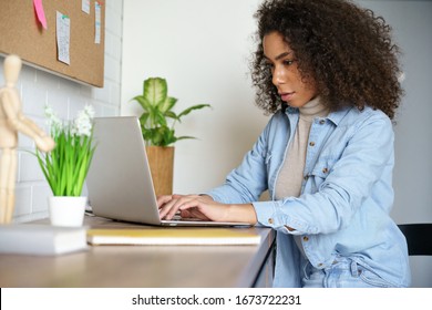 Focused Young African American Teen Girl College Student Using Laptop Computer Typing Studying Working Online. Serious Mixed Race Millennial Woman Doing Internet Research Sitting At Home Office Desk.