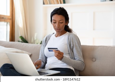 Focused Young African American Woman Holding Credit Bank Card, Entering Payment Information In Online Shopping Application, Purchasing Good Or Services In Internet Store Using Computer At Home.