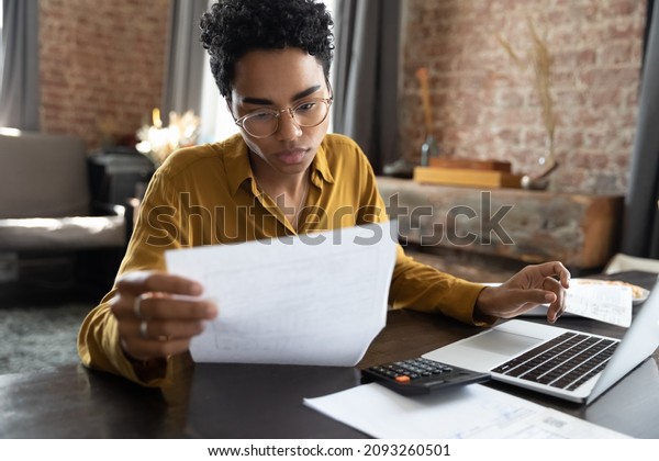 Focused young African American woman in\
eyeglasses looking through paper documents, managing business\
affairs, summarizing taxes, planning future investments, accounting\
alone at home office.