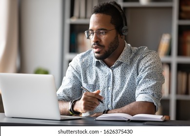 Focused young african businessman wear headphones study online watching webinar podcast laptop listening learning education course conference calling make notes sit at work desk  elearning concept