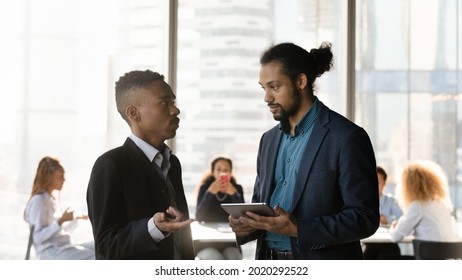 Focused Young African Ethnicity Businessman Consulting With Biracial Male Partner, Holding Digital Tablet In Hands. Concentrated Two Mixed Race Employees Colleagues Discussing Online Project Results.