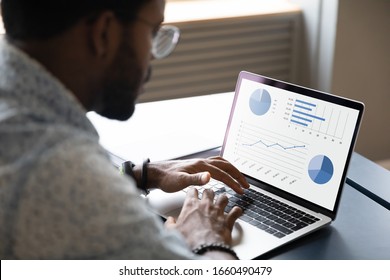 Focused young african ethnicity male marketing specialist professional working on project report, analyzing graphs charts in economic application on computer, sitting at table in office or home.