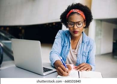 Focused young African female entrepreneur sitting at a table in a modern office building lobby working on a laptop and writing notes in her planner