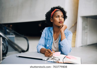 Focused young African female entrepreneur deep in thought while working at a table in a modern office building lobby - Shutterstock ID 703779808