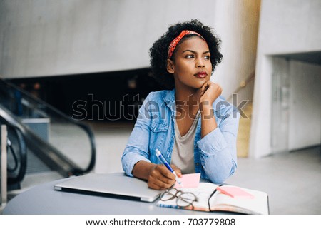 Focused young African female entrepreneur deep in thought while working at a table in a modern office building lobby