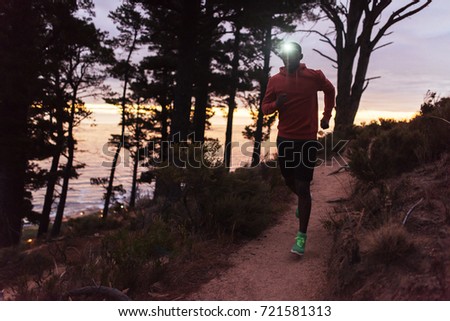Focused young African man wearing a headlamp running alone down a trail in the forest while out for a cross country run at dusk