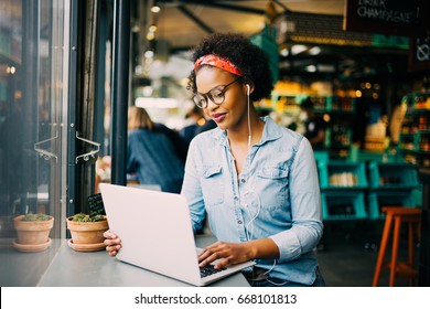 Focused young African woman sitting alone at a counter in a cafe working on a laptop and listening to music on earphones