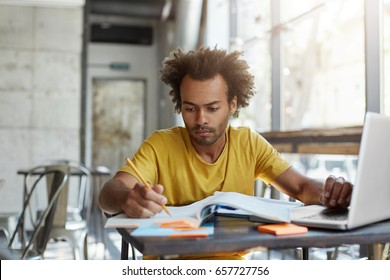 Focused Young Afro American English Teacher Checking His Students' Copybooks, Sitting At Cafe Table In Front Of Open Laptop Computer. Serious Black Male Student Learning Lesson At University Canteen