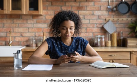 Focused Young Afro American Lady Sit At Kitchen Table Check Electronic Day Planner In Phone App Planning Meeting. Concentrated Black Woman Student Learning From Home Read Email Message On Cell Screen