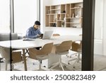 Focused young Arab employee man using digital device for business communication, sitting at large table alone in office co-working space, typing pn smartphone at laptop computer