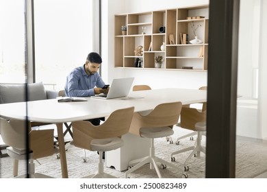 Focused young Arab employee man using digital device for business communication, sitting at large table alone in office co-working space, typing pn smartphone at laptop computer - Powered by Shutterstock