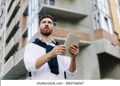 Focused young architect using digital tablet while standing at construction site. Bearded specialist with modern tablet controlling working process.