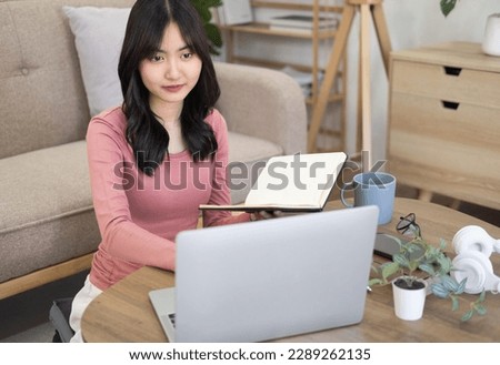 Focused young asian  businesswoman or student looking at laptop holding book learning, serious woman working or studying with computer doing research or preparing for exam online.