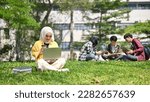 Focused young Asian Muslim female college student in hijab sitting on the grass, sipping coffee and using her laptop in the campus park. Uni-lifestyle concept