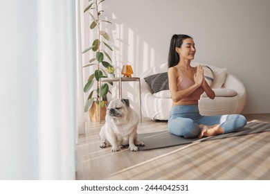 Focused young Asian woman enjoys a meditative yoga pose accompanied by her loyal pug dog in a bright, airy living space with soft morning light - Powered by Shutterstock