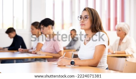 Focused young attractive Hispanic woman listening to lecture and taking notes in classroom with group of adult people. Postgraduate education concept