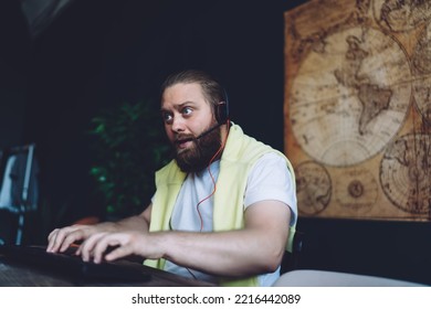 Focused Young Bearded Male Gamer In White T Shirt And Headset With Mic Using Keyboard While Playing Video Game On Computer Sitting On Chair At Desk At Home