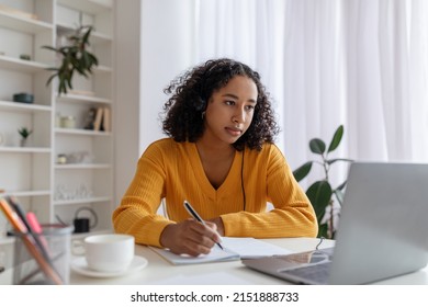 Focused Young Black Woman In Headset Having Online Lesson On Laptop, Writing In Notebook At Home. Serious African American Female Student Taking Notes During Webinar, Studying Remotely