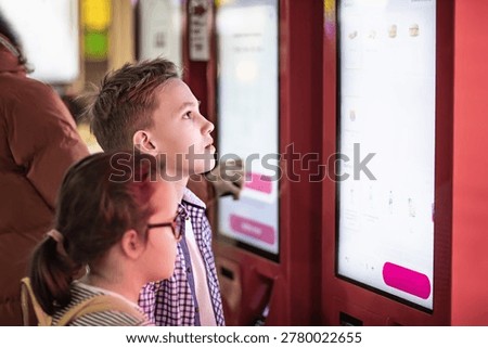 A focused young boy interacts with a red digital kiosk screen while another child observes. Fast food cafe.