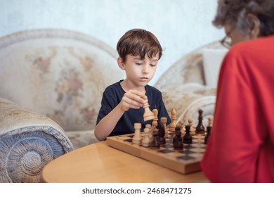 Focused young boy iplaying chess with an elderly woman in a cozy living room setting, highlighting intergenerational bonding and strategic thinking. Grandson and grandmother. International Chess Day. - Powered by Shutterstock