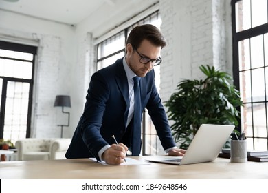 Focused Young Businessman In Formal Suit And Glasses Working On Computer, Standing At Table, Handwriting Notes In Paper Report. Busy 30s Employee Managing Tasks Or Doing Online Research Analysis.