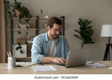Focused young businessman freelancer engaged in distant work from home office desk make report on laptop type document in app. Serious guy student in glasses prepare for exam using online literature
