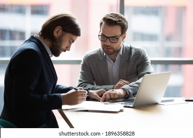 Focused young businessman signing agreement with skilled lawyer in eyeglasses. Concentrated financial advisor showing place for signature on paper contract document to male client at meeting in office