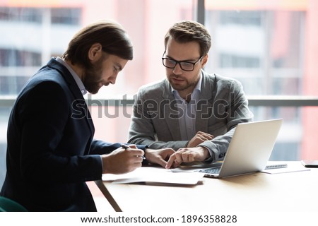 Focused young businessman signing agreement with skilled lawyer in eyeglasses. Concentrated financial advisor showing place for signature on paper contract document to male client at meeting in office 商業照片 © 