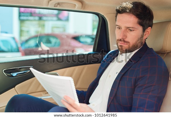 Focused\
young businessman sitting in the backseat of a car driving through\
the city reading paperwork and using a\
cellphone