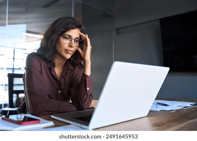 Focused young businesswoman looking at laptop pc computer screen having headache, migraine. Latin business woman holding hand near temples, feeling stressed and tired sitting at workplace in office - Powered by Shutterstock