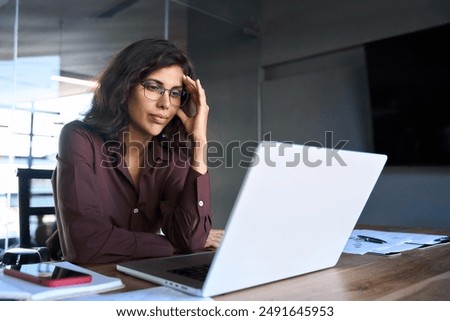 Focused young businesswoman looking at laptop pc computer screen having headache, migraine. Latin business woman holding hand near temples, feeling stressed and tired sitting at workplace in office