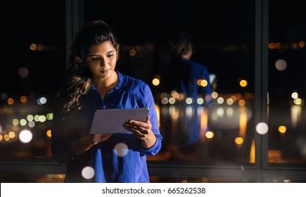 Focused young businesswoman using a digital tablet while standing alone in an office late at night in front of windows overlooking the city  - Powered by Shutterstock