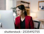 Focused young businesswoman works late at night in her office, browsing, searching, and typing on her computer screen in a stylish red blazer, showing determination and concentration