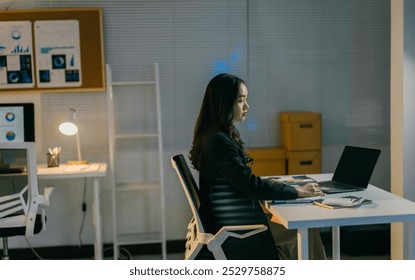 Focused young businesswoman works late at her office desk, using a laptop to complete a project in a dimly lit room - Powered by Shutterstock