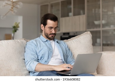 Focused Young Caucasian Man In Glasses Sit On Sofa At Home Work Online On Laptop Gadget. Concentrated Millennial Male Typing On Computer Browse Wireless Internet On Device. Technology Concept.