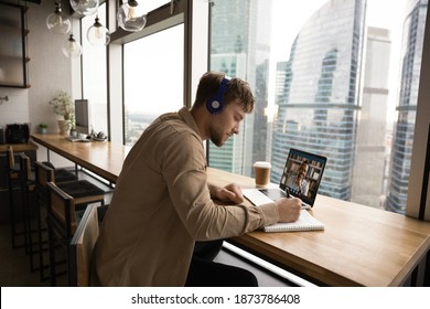 Focused Young Caucasian Man In Headphones Sit At Desk Make Notes Study Distant Talk On Video Call On Laptop. Millennial Male In Earphones Have Webcam Digital Virtual Conference Or Event On Computer.