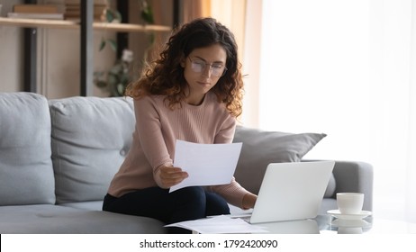 Focused young Caucasian woman in glasses sit on couch at desk work on laptop read paperwork, concentrated millennial businesswoman manage paper documents, pay bills or taxes, use computer at home