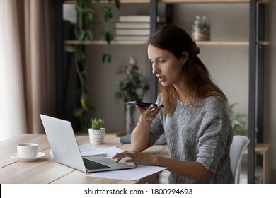 Focused young Caucasian woman sit at desk at home work on laptop record audio message on cellphone, millennial female use computer browsing, activate virtual digital voice assistant on smartphone - Powered by Shutterstock