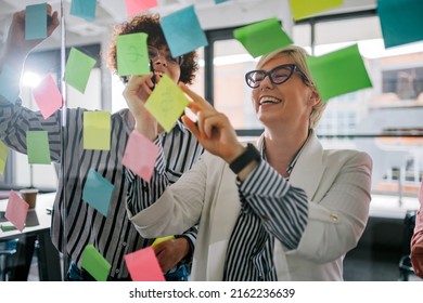 Focused young diverse teammates and their team leader working on an IT startup project and discussing details and tasks while writing notes on colorful paper stickers on a glass wall in the office. - Powered by Shutterstock