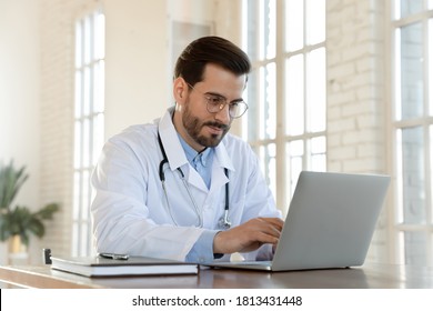 Focused Young Doctor Wearing Glasses Working On Laptop In Office, Sitting At Desk, Looking At Screen, Serious Therapist Gp Writing Report Or Email, Consulting Patient Online, Filling Medical Card